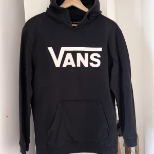 Svart hoodie från Vans st Junior XL - Säljer en svart hoodie från Vans med deras ikoniska logga i vitt på framsidan. Tröjan har en stor ficka framtill och en bekväm huva. Perfekt för en avslappnad stil. Ingen anmärkning. Inga hål, fläckar eller dylikt.