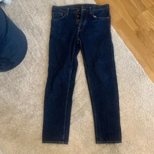 Mörkblå jeans från Nudie Jeans - Snygga mörkblå jeans från Nudie Jeans med knappgylf och klassisk femficksdesign. Perfekta för en stilren look. Passar både till vardags och mer uppklädda tillfällen. Regular-fit jeans med avsmalnande ben i djup mörk indigo-tvätt med subtila skavsår och slitage. Tillverkad av ekologisk bomull.