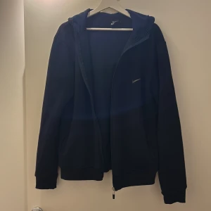 Svart hoodie från Nike - Säljer en svart hoodie från Nike med dragkedja och huva. Den har en diskret Nike-logga på bröstet och är perfekt för en avslappnad stil. Passar bra till både träning och vardag.