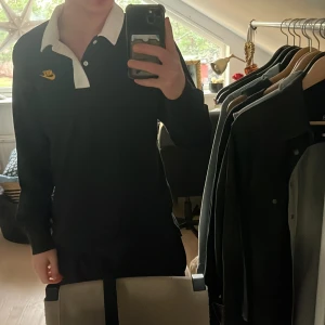 Svart pikétröja från Nike - Säljer en svart pikétröja från Nike med vit krage och knappar. Tröjan har ett gult Nike-märke på bröstet och en stor logga på ryggen. Perfekt för en sportig look!