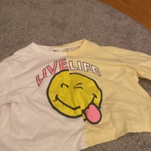 Gul och vit t-shirt från H&M - Cool t-shirt från H&M i samarbete med Smiley World. Den är delad i gult och vitt med ett stort smileytryck och texten 'LIVE LIFE' i rosa och vitt. Perfekt för lite yngre barn.