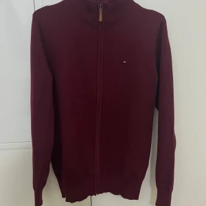 Vinröd kofta med dragkedja från Tommy Hilfiger - Snygg vinröd kofta från Tommy Hilfiger med dragkedja och liten logga på bröstet. Perfekt för kyliga dagar och enkel att matcha med andra plagg.  Tröjan äe storlek M men passar också som S