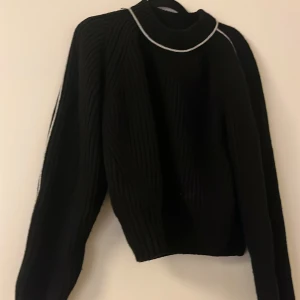 Svart stickad tröja med vita detaljer - Snygg svart stickad tröja med vita detaljer runt halsen och längs ärmarna. Perfekt för kyliga dagar med sin höga krage och långa ärmar. En stilren och bekväm tröja för alla tillfällen.