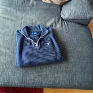 Blå ribbad tröja från Gant - Säljer en stilren blå ribbad tröja från Gant med dragkedja vid halsen. Tröjan har en klassisk design med en liten logotyp på bröstet. Perfekt för en avslappnad och snygg look. Riktigt snyggt nu till hösten