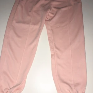 Rosa mjukisbyxor från Nike - Säljer ett par bekväma rosa mjukisbyxor från Nike. Perfekta för en avslappnad stil med elastisk midja och ribbade muddar vid bensluten. Byxorna har en diskret Nike-logga på höften.