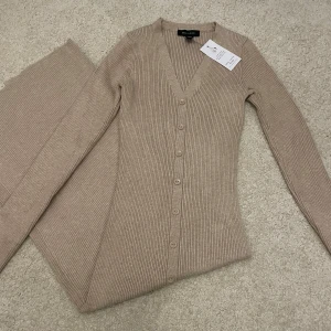 Beige ribbad/stickad klänning från New Look - Snygg beige ribbad klänning från New Look med v-ringning och knappar framtill. Klänningen har långa ärmar och en slits som ger extra stil. Perfekt för både vardag och speciella tillfällen.