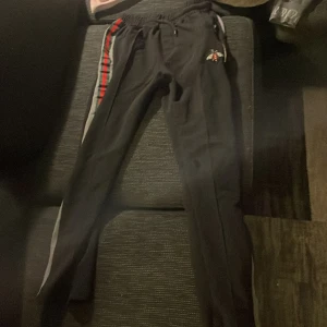 Svarta byxor med broderad detalj - Snygga svarta byxor med en broderad bi-detalj på sidan. Byxorna har en röd och grön rand längs benen och dragkedjor vid fickorna. Perfekta för en avslappnad stil.