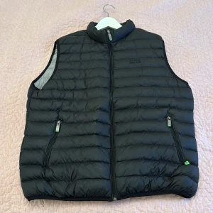 Boss - Snygg svart dunväst från Hugo Boss med dragkedja och broderad logga på bröstet. Perfekt för kyligare dagar med sin quiltade design och praktiska fickor med dragkedja.