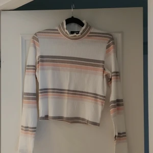 Randig ribbad tröja från BikBok - Säljer en snygg randig tröja från BikBok i storlek M. Tröjan har en ribbad struktur med långa ärmar och en hög krage. Färgerna är en mix av vitt, beige och rosa, vilket ger en stilren look. Perfekt för både vardag och speciella tillfällen.
