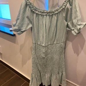 Ljusgrön klänning med volangdetaljer - Säljer en superfin ljusgrön klänning med volangdetaljer. Den har en offshoulder-stil med korta ärmar och en smockad midja som ger en snygg passform. Perfekt för sommarens alla fester eller en mysig dag på stan!