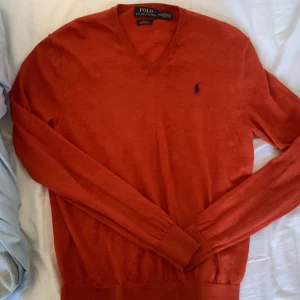 Orange tröja från Polo Ralph Lauren - Säljer en snygg orange tröja från Polo Ralph Lauren i merinoull. Den har en klassisk v-ringning och är långärmad. Perfekt för höst och vinter, och den har den ikoniska loggan broderad på bröstet. Supermjuk och stilren!