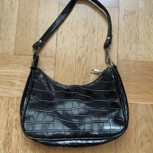 Svart axelväska med krokodilmönster - Säljer en svart axelväska med krokodilmönster. Väskan har en glansig finish och en dragkedja upptill. Skriv gärna om ni har frågor 💕
