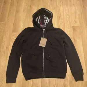 Svart huvtröja från Burberry - Säljer en stilren svart huvtröja från Burberry med dragkedja och klassiskt rutigt foder i huvan. Tröjan har en broderad logga på bröstet och ribbade muddar vid ärmslut och nederkant. Perfekt för en casual look! Har storlek S och L.