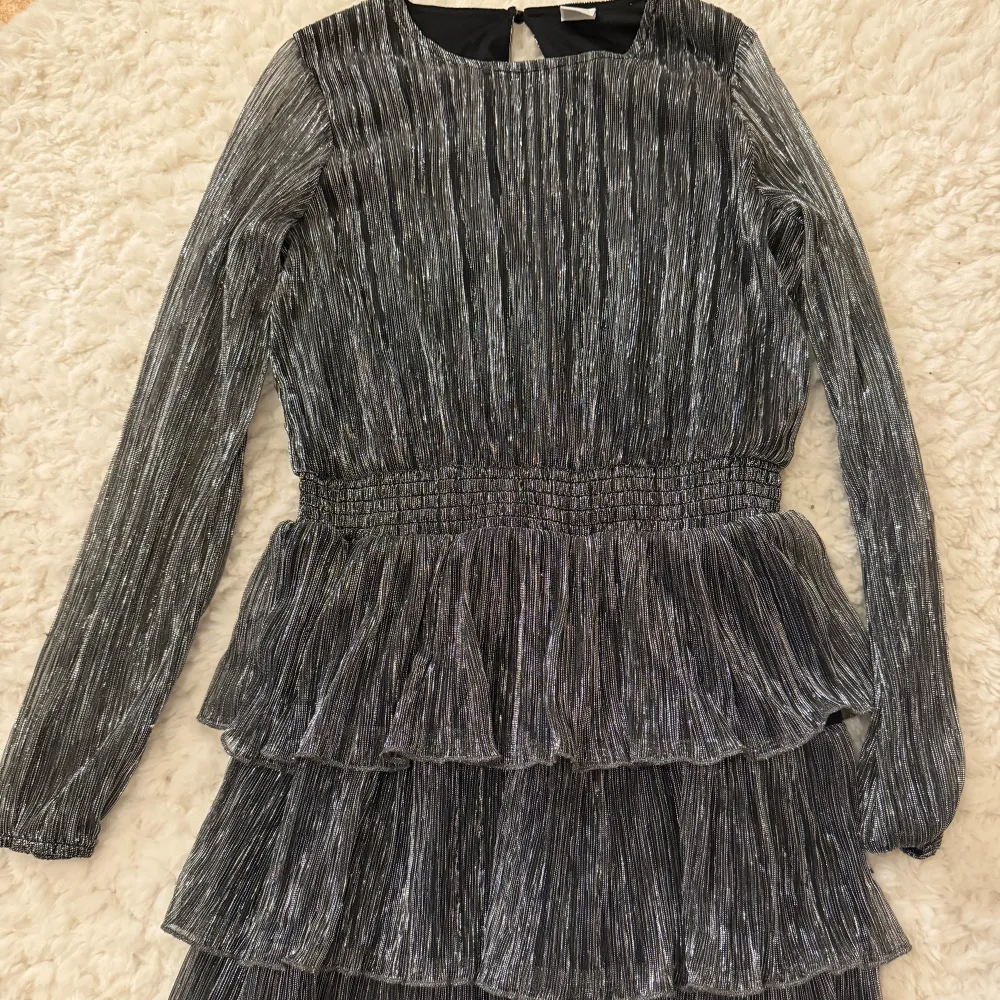 Säljer en glittrig klänning från Lindex i en snygg silverfärg. Klänningen har långa ärmar och flera lager med volanger som ger en festlig look.Jag har använt den förra året på nyår sedan så hade jag växt ur den 🩷. Mekot.