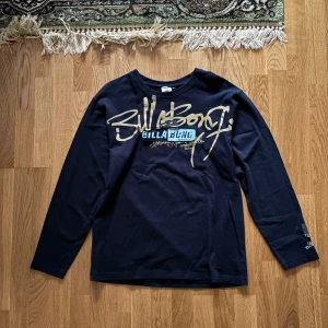 Svart långärmad tröja från Billabong - Säljer en svart långärmad tröja från Billabong med ett stort tryck i guld och blått på framsidan. Perfekt för en avslappnad stil. Tröjan har en rund halsringning och är gjord i ett mjukt material.