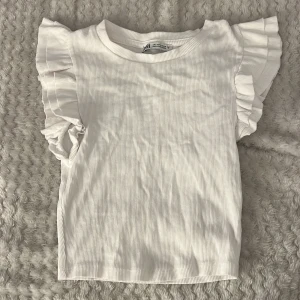 Vit ribbad topp från Zara - Säljer en söt vit ribbad topp från Zara med volangdetaljer på ärmarna. Perfekt för en stilren och chic look. Toppen har en rund halsringning och korta ärmar med volanger som ger en extra touch.