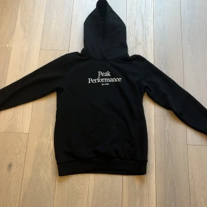Svart hoodie från Peak Performance - Säljer en stilren svart hoodie från Peak Performance med vit logga på bröstet. Tröjan har en klassisk känguruficka och en bekväm huva. Perfekt för en avslappnad look. hoodien är knappt använd och inga tecken på skador eller slitage så hör av dig om du har några funderingar 👍