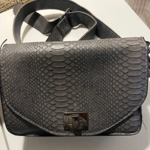 Grå axelväska med ormskinnsmönster - Snygg grå axelväska med ormskinnsmönster och justerbar axelrem. Väskan har ett stilrent metallspänne framtill och gott om plats inuti för dina nödvändigheter. Perfekt för att ge din outfit en trendig touch. Köpt för 150kr säljs för 75kr