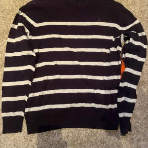 Randig tröja från Tommy Hilfiger - Säljer en snygg randig tröja från Tommy Hilfiger i svart och grått. Tröjan har långa ärmar och en klassisk rund halsringning. Perfekt för en stilren look.