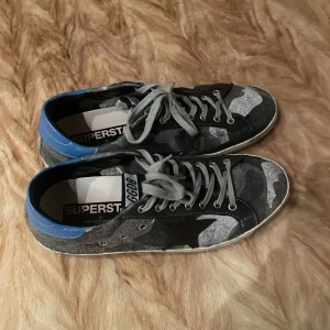 Camouflage sneakers med blå detaljer - Goolden goose superstar  sneakers med ett coolt camouflage-mönster i grå och svart. Skorna har blå detaljer vid hälen och grå snörning. Perfekta för en avslappnad stil.