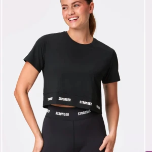 Svart croppad t-shirt från Stronger - Snygg svart croppad t-shirt från Stronger med korta ärmar och elastisk kant med logotyp. Perfekt för en sportig look eller träning. Nypris 499.