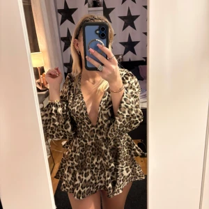 Leopardmönstrad klänning med volang - Trendig leopardmönstrad klänning med volangdetaljer och djup v-ringning. Klänningen har långa ärmar och en kort längd som ger en chic look. Perfekt för att sticka ut med stil.