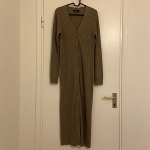 Beige ribbad långklänning från KappAhl - Snygg mörk beige långklänning från KappAhl i ribbat material. Klänningen har en v-ringad design med knappar längs hela framsidan och långa ärmar. Den kan även var en lång kofta om man har den öppen. Perfekt för en stilren look.