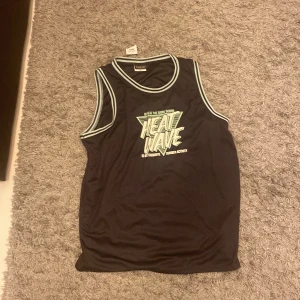 Svart linnetopp från Alphar One - Säljer en svart linnetopp från Alphar One med trycket 'Heat Wave' i neonfärger på framsidan. Toppen har vita kantband runt ärmhål och halsringning. Perfekt för varma dagar eller träning.