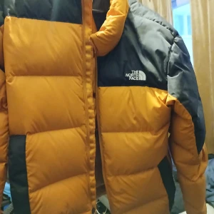 Orange och svart dunjacka från The North Face - Tar bara kontant möts i Eskilstuna.Säljer en snygg och varm dunjacka från The North Face i orange och svart. Jackan har en dragkedja och långa ärmar, perfekt för kalla dagar. Den har en klassisk pufferdesign med logotyp på bröstet. Inuti finns en praktisk ficka med dragkedja.
