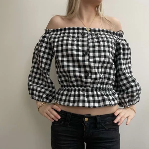 Svartvit rutig offshoulder blus - Säljer en trendig svartvit rutig offshoulder blus med knappar framtill. Blusen har trekvartsärmar och en elastisk midja som ger en fin passform. Perfekt för en stilren look!