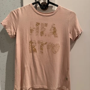 Rosa t-shirt från Pomp de Lux - Säljer en söt rosa t-shirt från Pomp de Lux med ett glittrigt mönster framtill. T-shirten har korta ärmar och en rund halsringning. Perfekt för en avslappnad stil. Strl 134/140, pris 35kr. Aldrig använd