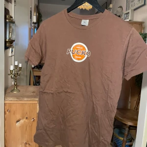 Brun t-shirt med Pucko-tryck - Säljer en brun t-shirt med ett coolt Pucko-tryck i orange och vitt på framsidan. T-shirten har en klassisk rund hals och korta ärmar. Perfekt för dig som gillar retrostil och choklad!
