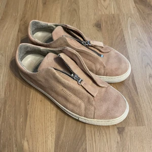 Ljusrosa/Beige Axel Arigatos i Mocka med zip - Snygga beige sneakers i mocka från Axel Arigato med dragkedja framtill. Skorna har en stilren design med vit sula och är perfekta för en avslappnad look. Nyåris 2300kr