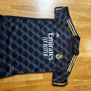 Real Madrid Tröja - ÄKTA, S men passar M                                                                           Snygg svart fotbollströja från Adidas med Real Madrid-logga på bröstet. Tröjan har ett unikt mönster och korta ärmar med gula detaljer. Perfekt för alla fotbollsfans som vill visa sitt stöd för laget.