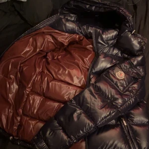 Murk blå pufferjacka från Moncler - Säljer en snygg murkblå pufferjacka från Moncler med en glansig finish. Jackan har en hög krage och dragkedja framtill. Insidan är fodrad i en mörkröd nyans som ger en cool kontrast. Perfekt för kyliga dagar! 🖤TAR BA BYTE!! Elr möts upp