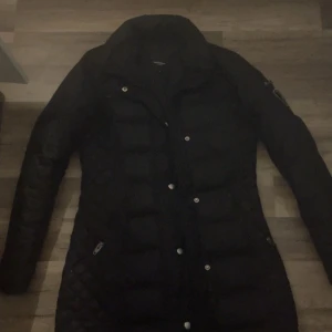 Svart dunjacka från Rockandblue - Snygg svart dunjacka från Rockandblue med dragkedja och knappar. Perfekt för kyliga dagar med sin långa design och quiltade detaljer. Jackan har en stilren look med Rockandblue-logga på ärmen.