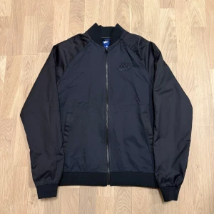 Mörkblå/svart bomberjacka från Nike - Säljer en stilren svart bomberjacka från Nike. Jackan har en dragkedja framtill och ribbade muddar vid ärmslut och nederkant. Perfekt för en sportig look. Pris kan diskuteras.