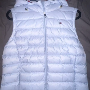 Vit dunväst från Tommy Hilfiger - Snygg vit dunväst från Tommy Hilfiger med dragkedja och huva. Västen har en klassisk design med märkets logga på bröstet. STORLEK XS MEN STÖRRE ÄN VAD DET SER UT!