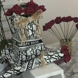  Buket från Dior med dekorativa rosor - Elegant parfym från Dior, vackert inslagen med ett mönstrat omslagspapper och dekorerad med röda rosor. Paketet pryds av en lyxig guldfärgad rosett med pärldetaljer. Perfekt som en exklusiv present.