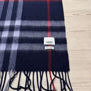Burberry halsduk - Säljer en klassisk rutig halsduk från Burberry i kashmir. Den har en elegant kombination av svart, grått och rött med fransar i ändarna. Perfekt för att hålla dig varm och stilren under kyliga dagar. | Skick: 10/10 (aldrig använd) | Urspungspris: 5825kr | Vårt pris: 1999kr | Tveka inte att skriva vid eventuella frågor! Mvh