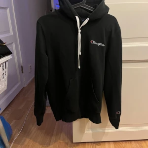 Svart hoodie från Champion - Säljer en klassisk svart hoodie från Champion med vit dragsko och broderad logga på bröstet. Tröjan har en ficka och långa ärmar. Perfekt för en avslappnad stil.