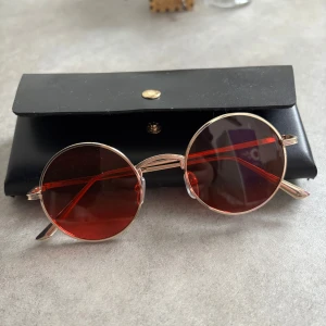 Runda solglasögon - Snygga runda solglasögon med rödtonade glas och en tunn metallbåge. Perfekta för att ge en retro touch till din stil. Kommer med ett svart fodral för säker förvaring.