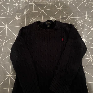 Mörkblå  stickad tröja från Ralph Lauren - Säljer en stilren mörkblå stickad tröja från Ralph Lauren med kabelstickat mönster. Tröjan har långa ärmar och en liten röd logga på bröstet. Perfekt för kyligare dagar!