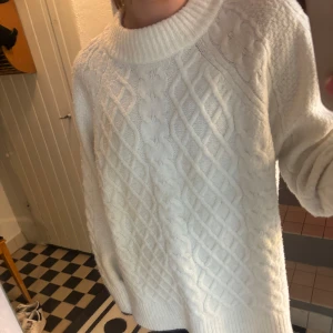 Vit stickad tröja - Säljer en mysig vit stickad tröja med ett klassiskt flätmönster. Tröjan har en hög krage och långa ärmar, perfekt för kyliga dagar. Den är både stilren och bekväm, ett måste i garderoben!