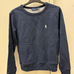 Blå tröja från Polo Ralph Lauren - Säljer en snygg blå tröja från Polo Ralph Lauren med deras ikoniska logga broderad på bröstet. Tröjan har långa ärmar och en rund halsringning, perfekt för en stilren look. Passar bra till både jeans och chinos.