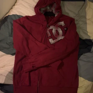 Vinröd hoodie från DC - Säljer en vinröd hoodie från DC med dragkedja och stort logotryck på bröstet. Perfekt för en avslappnad stil med sin bekväma passform och huva. Ett måste för streetwear-entusiaster!