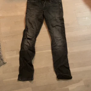 Svarta/gråa jeans från Jack & Jones - Snygga svarta jeans från Jack & Jones i modellen Slim/Glenn. De har en slim passform och är tillverkade i denim. Byxorna har slitna detaljer på benen för en trendig look.