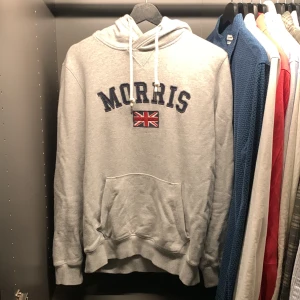 Grå hoodie från Morris - Säljer en snygg grå hoodie från Morris med broderad text och brittisk flagga på bröstet. Tröjan har en klassisk känguruficka och justerbar huva med snören. Perfekt för en avslappnad stil.