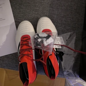 Rödvita fotbollsskor med dobbar - Säljer ett par snygga fotbollsskor i röd och vit färg med svarta detaljer. Skorna har snörning och är utrustade med dobbar för bra grepp på planen. Perfekta för fotbollsträning eller match!