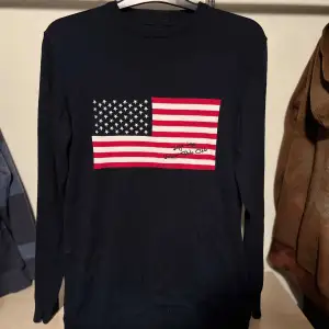 Säljer en snygg mörkblå tröja från U.S. Polo Assn. med en stor amerikansk flagga på framsidan. Tröjan har långa ärmar och är perfekt för en casual look. Passar bra till både jeans och chinos!